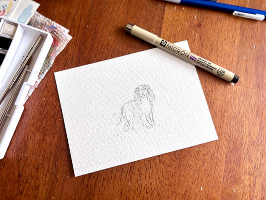 White watercolor paper with a dog drawing and a micron pen on a wooden surface