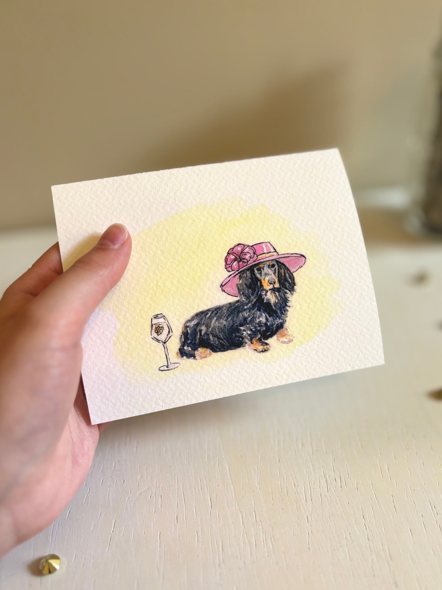 Hand holding an art print with a dachshund wearing a pink hat and glass of wine illustration.