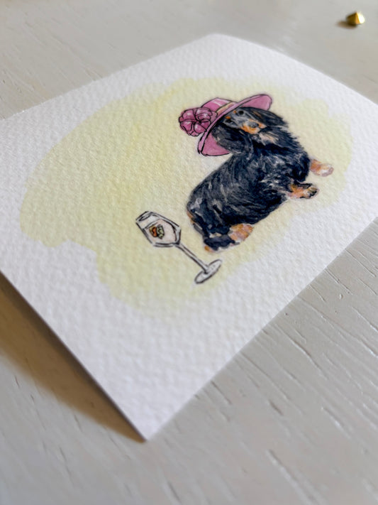 Watercolor art print with a dachshund wearing a pink hat and holding a glass of wine on a light wooden surface.
