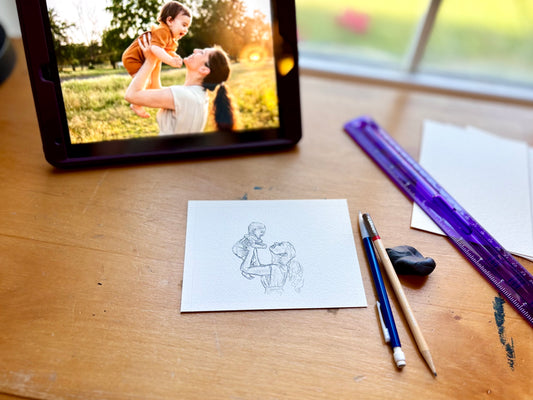 Tablet displaying a photo of a mother and child, with a drawing on paper and art supplies on a wooden surface.