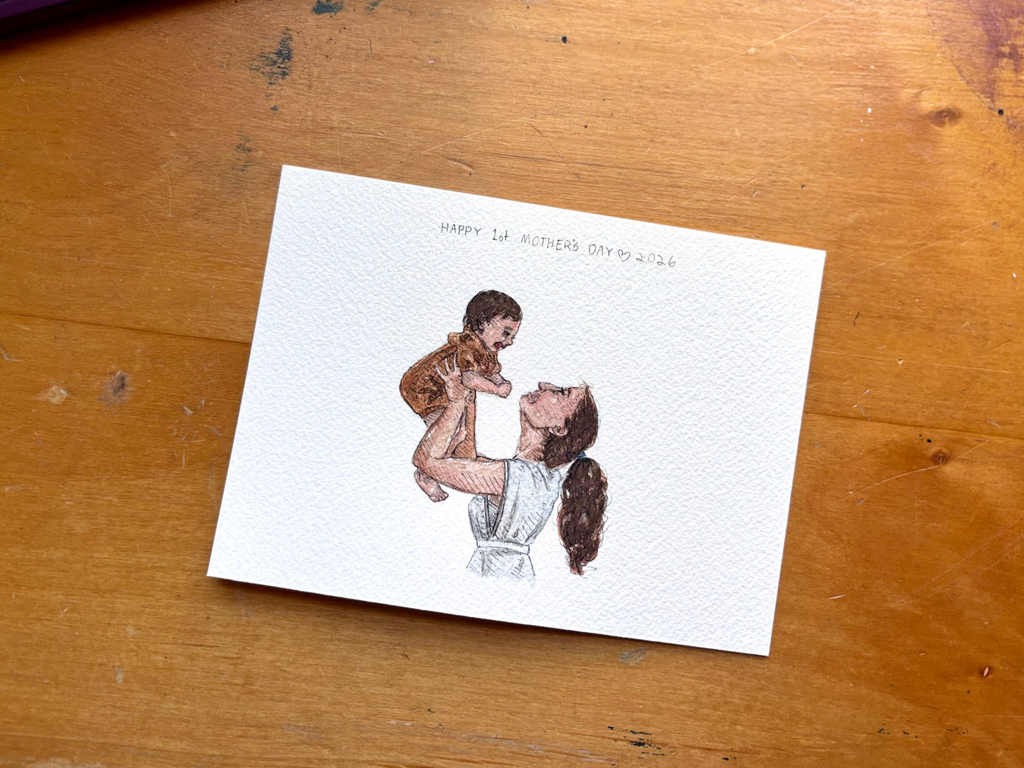 Illustrated Mother's Day card with a woman holding a child on a wooden surface.
