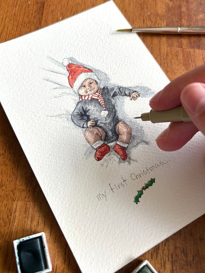 A close-up of a hand holding a fine-tip pen over a watercolor painting of a baby wearing a Santa hat, with the text 'my first Christmas.
