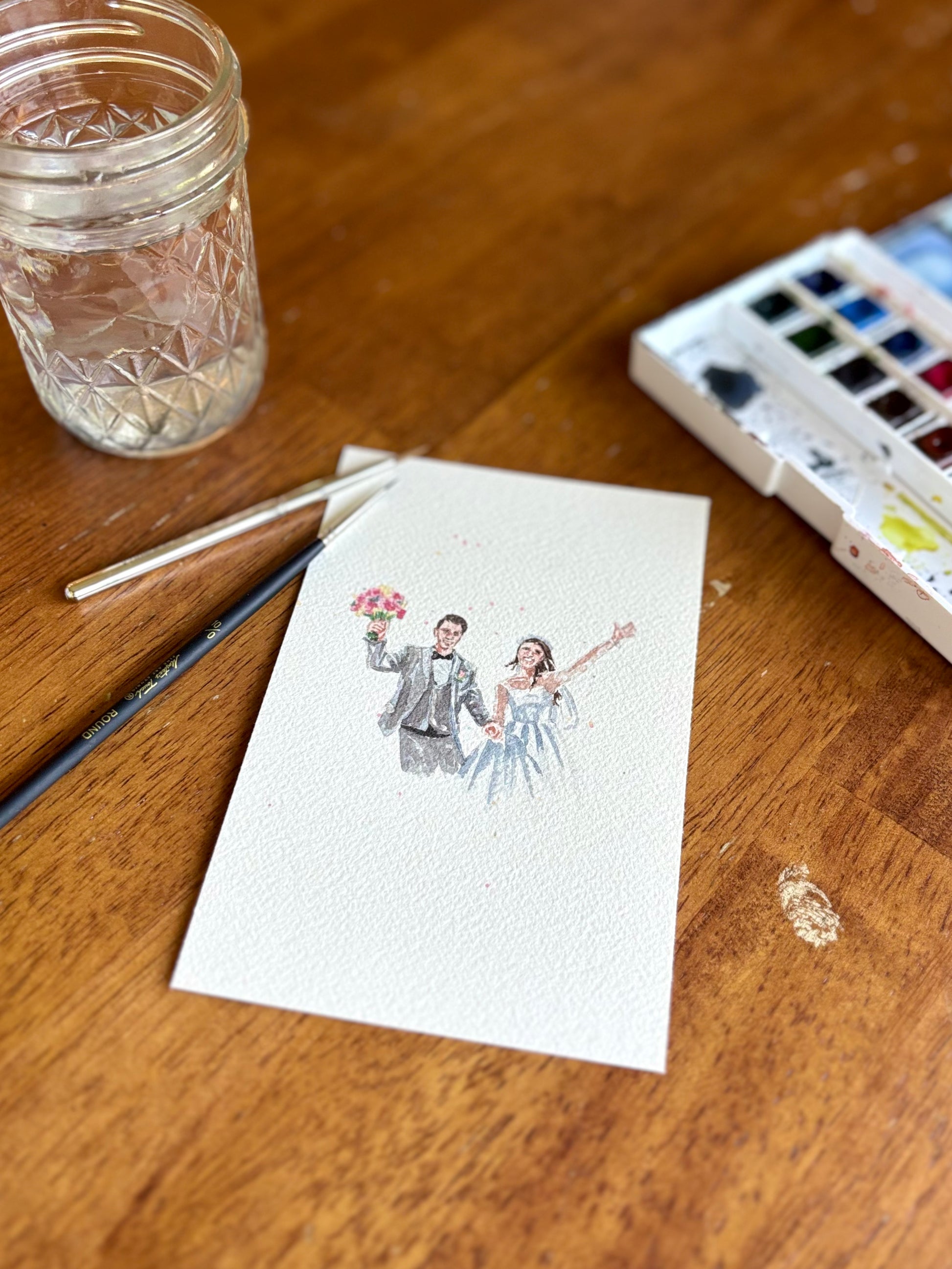 A small watercolor painting depicting a bride and groom, displayed on a wooden table alongside a paintbrush, a glass jar, and a watercolor palette.