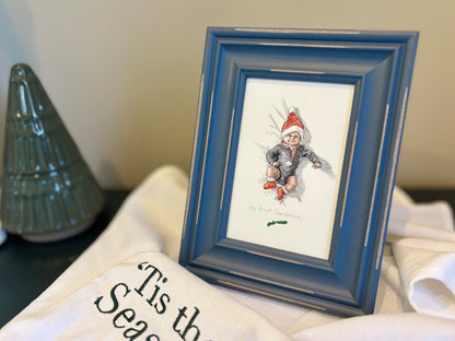 A blue wooden picture frame holding a vintage-style Christmas watercolor painting of a baby in a Santa hat, styled on a white fabric surface next to a green Christmas tree ceramic ornament.