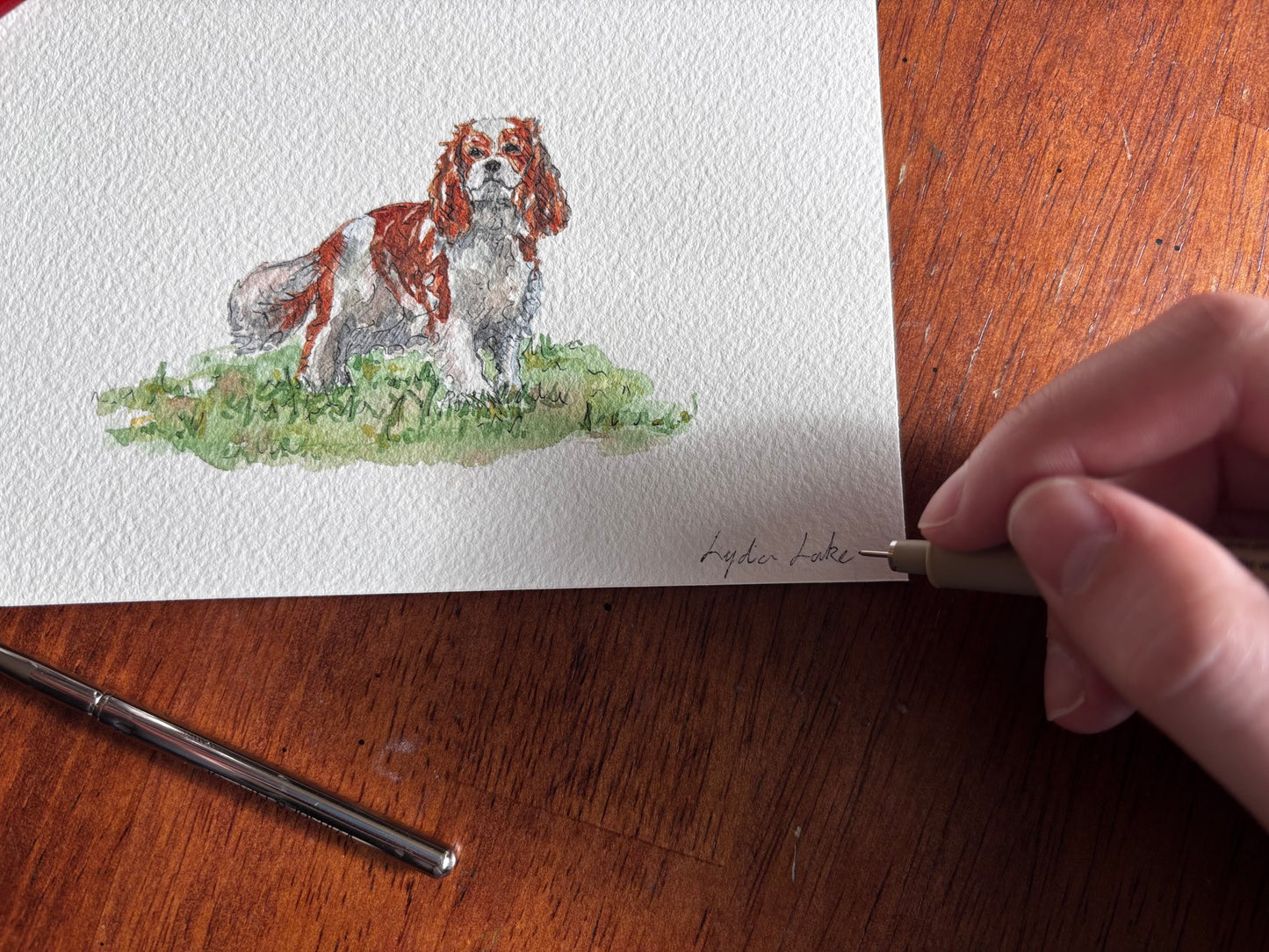 Hand signing an original watercolor portrait of a dog on a wooden surface