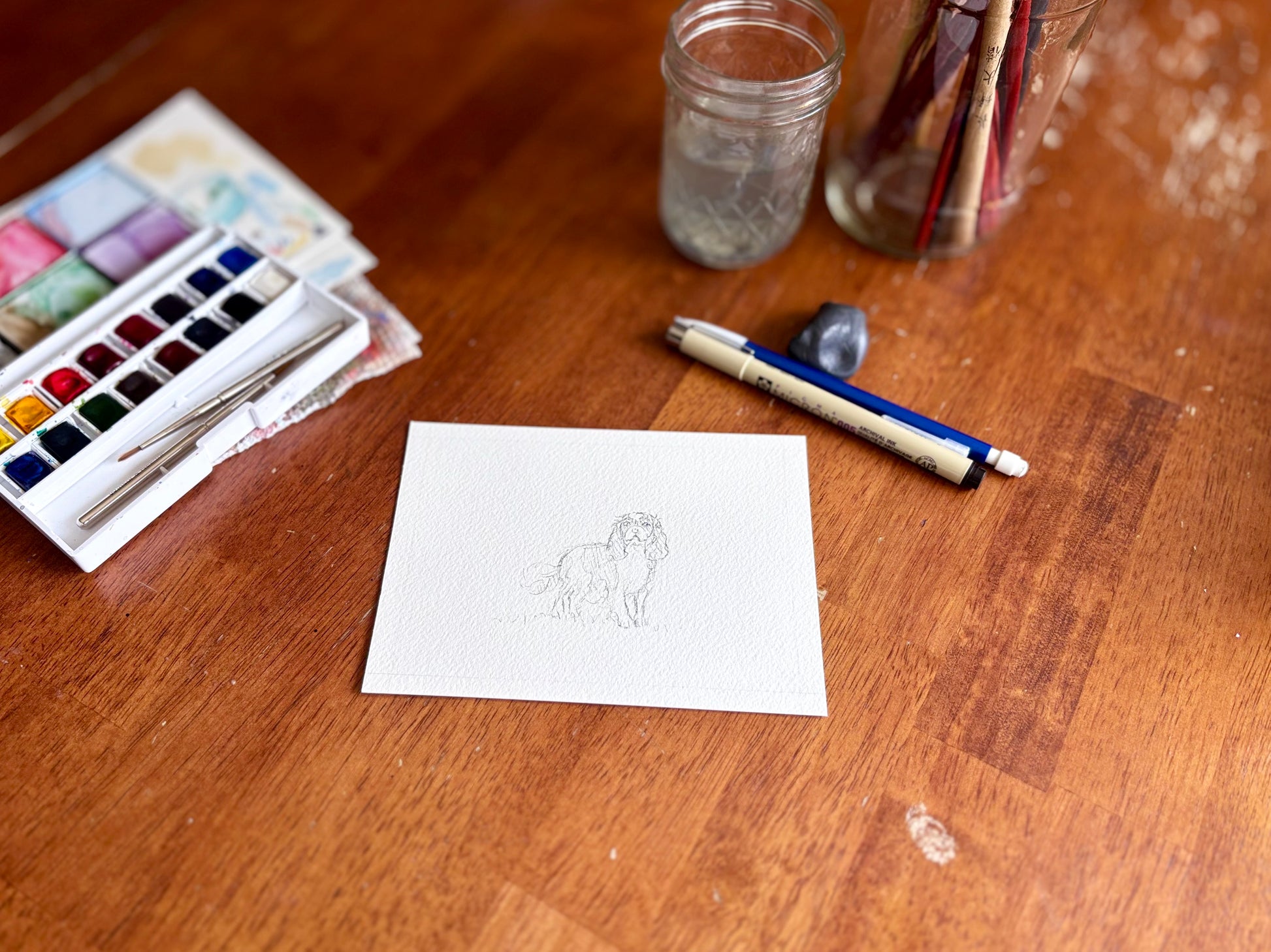 Art supplies including watercolor paints, a pen, and a small drawing on a wooden table.