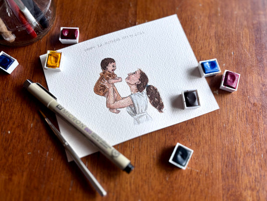 Aesthetically arranged watercolor paint and pens with a mother and son portrait.