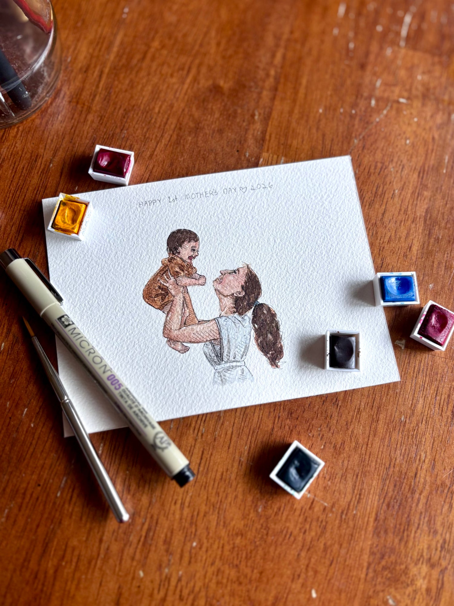 Watercolor illustration of a woman holding a child on a piece of paper with watercolor paint pans and a pen on a wooden surface.