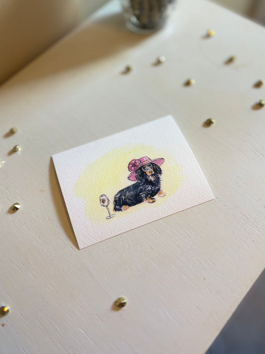 Miniature watercolor art print with a dog illustration wearing a hat on a light surface.