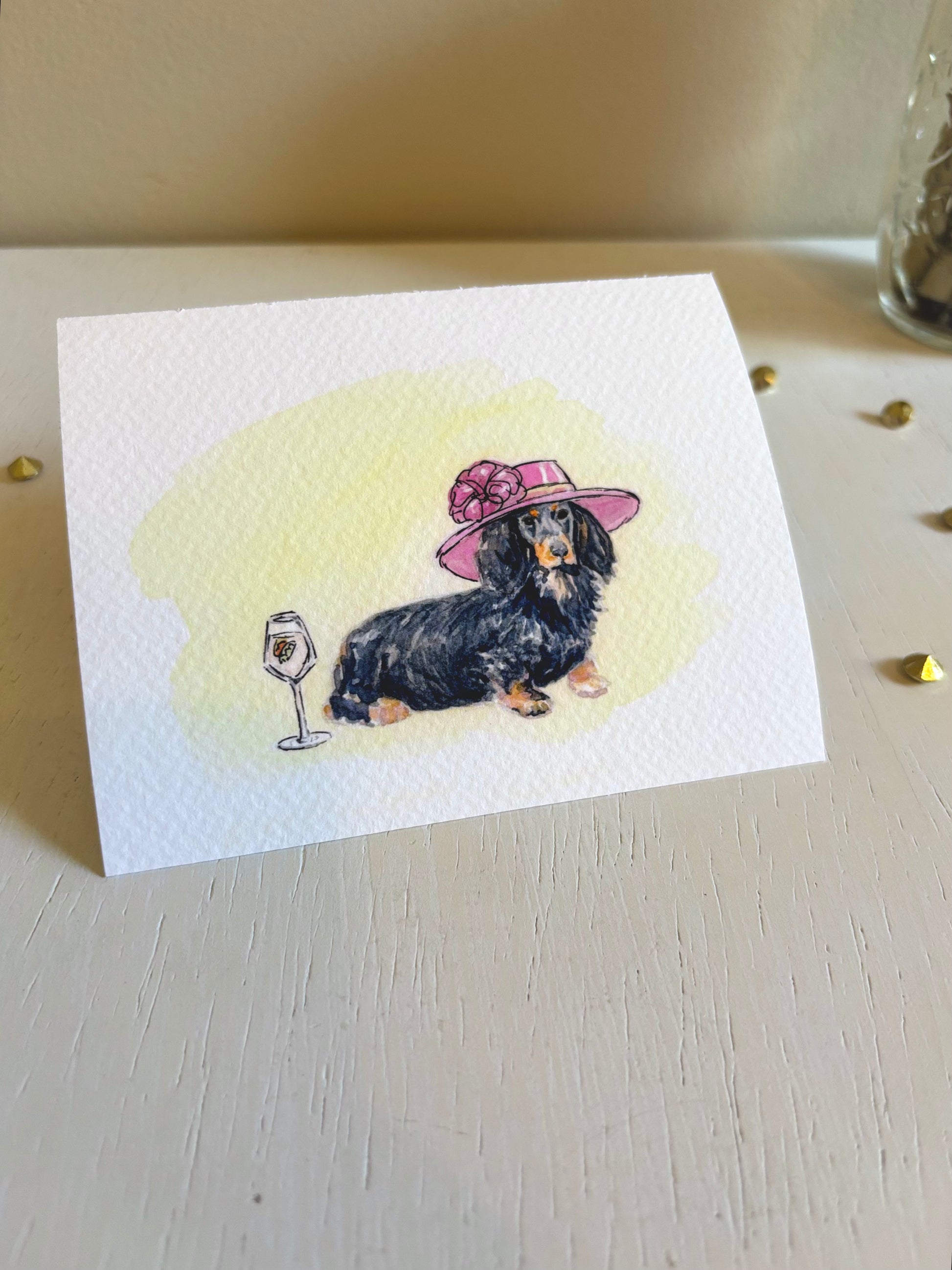 Miniature watercolor art print with a dachshund wearing a pink hat and a glass of wine on a light surface.