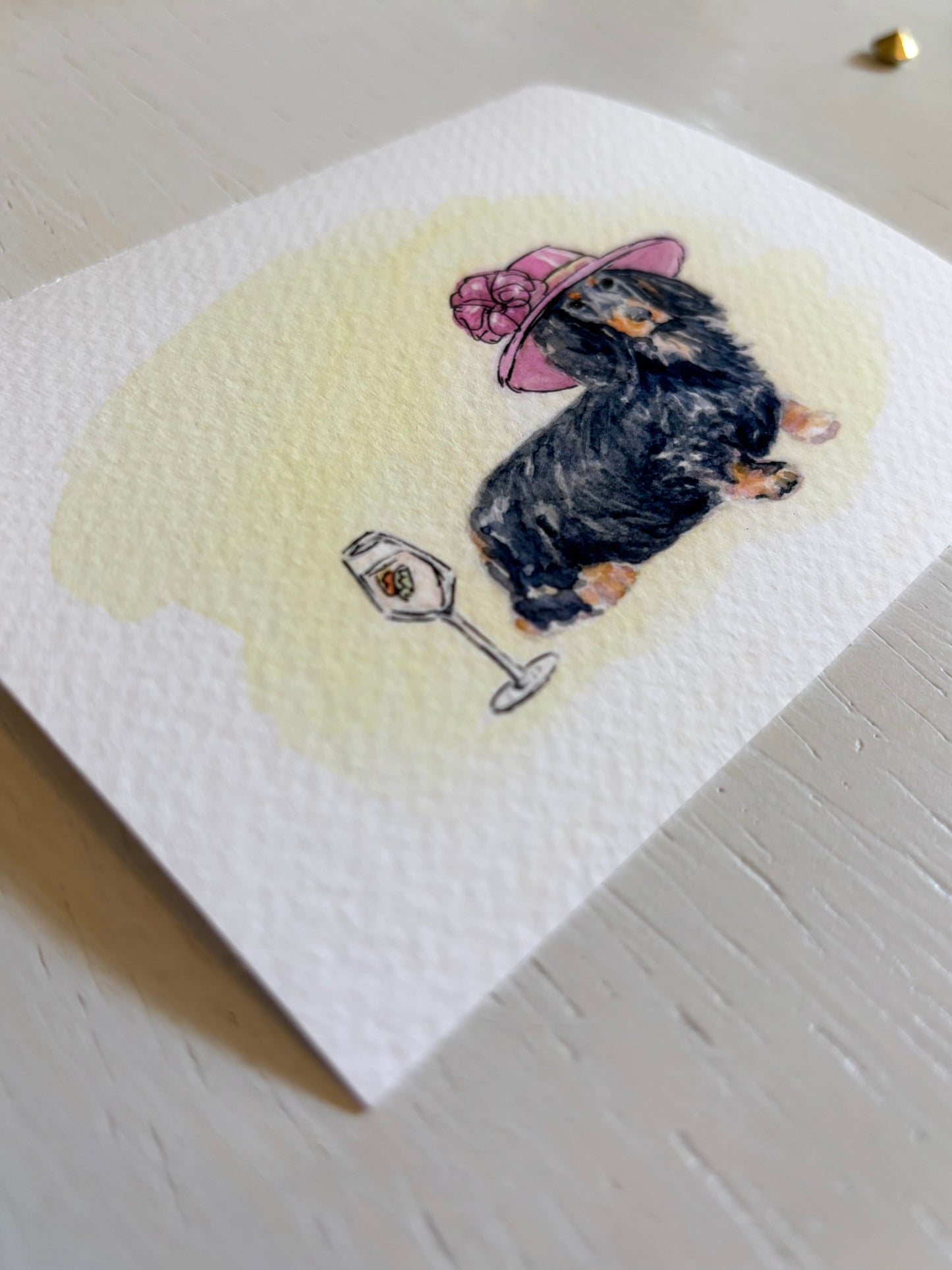 Watercolor art print with a dachshund wearing a pink hat and holding a glass of wine on a light wooden surface.