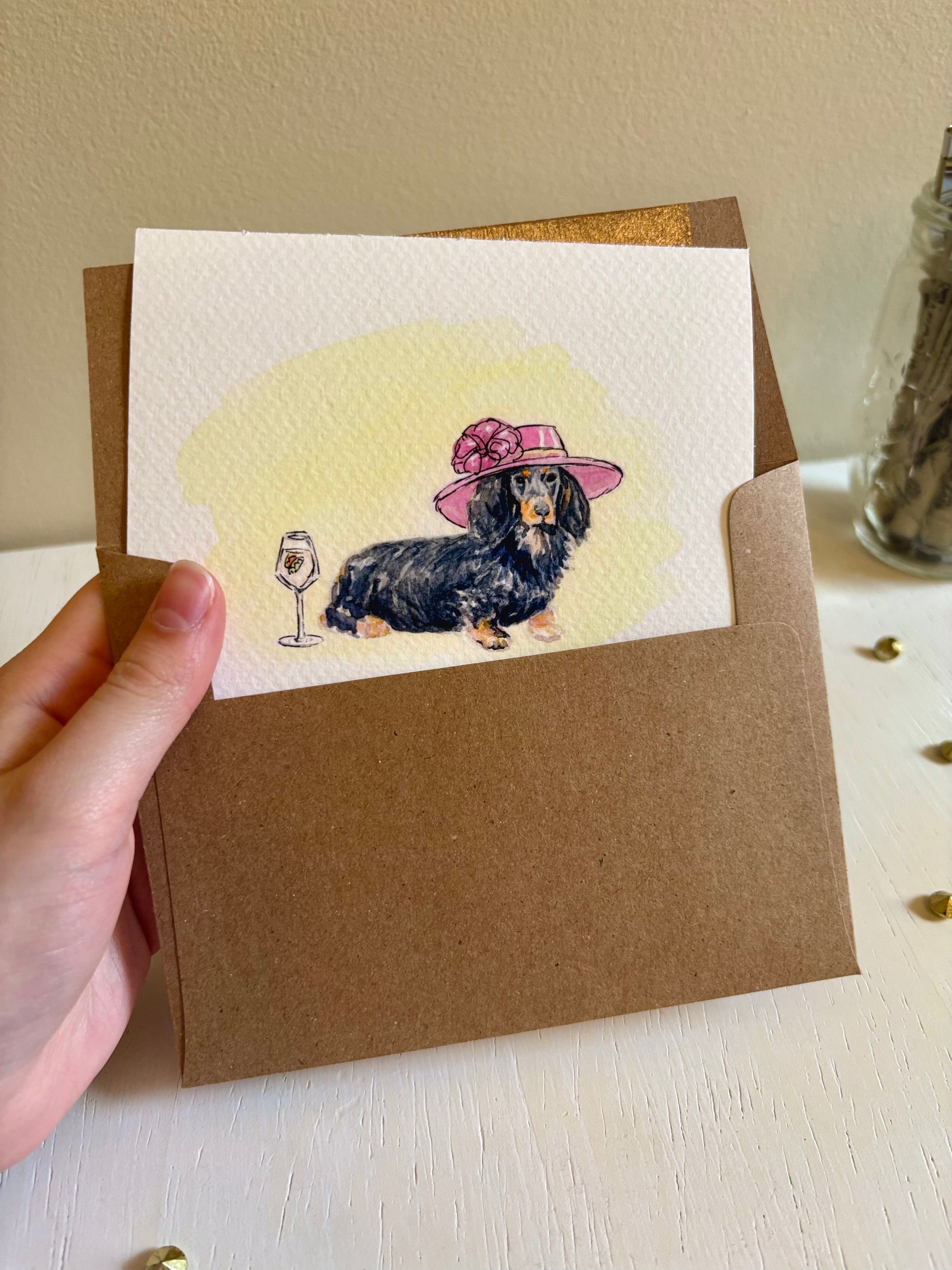 Hand holding a card with a dachshund illustration wearing a pink hat and a glass of wine in a brown envelope. 