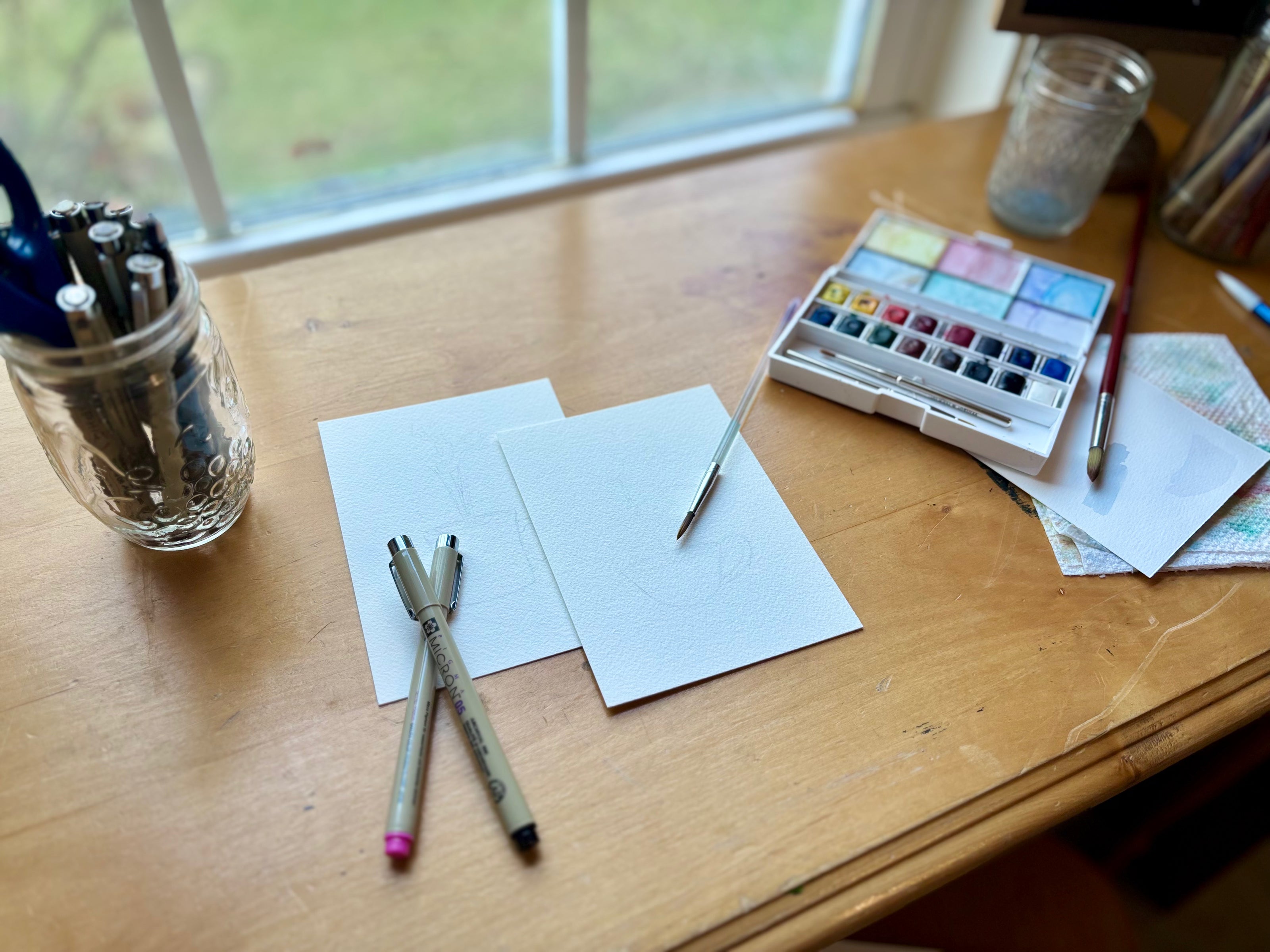 Art supplies including pens, watercolor paint, and brushes on a wooden table near a window.