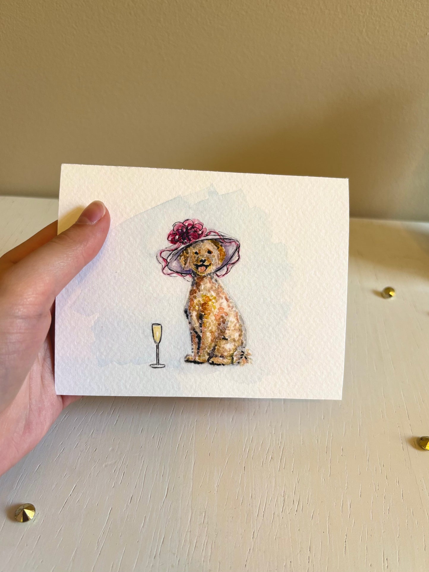 Hand holding a card with a Golden Doodle illustration wearing a hat on a light surface.