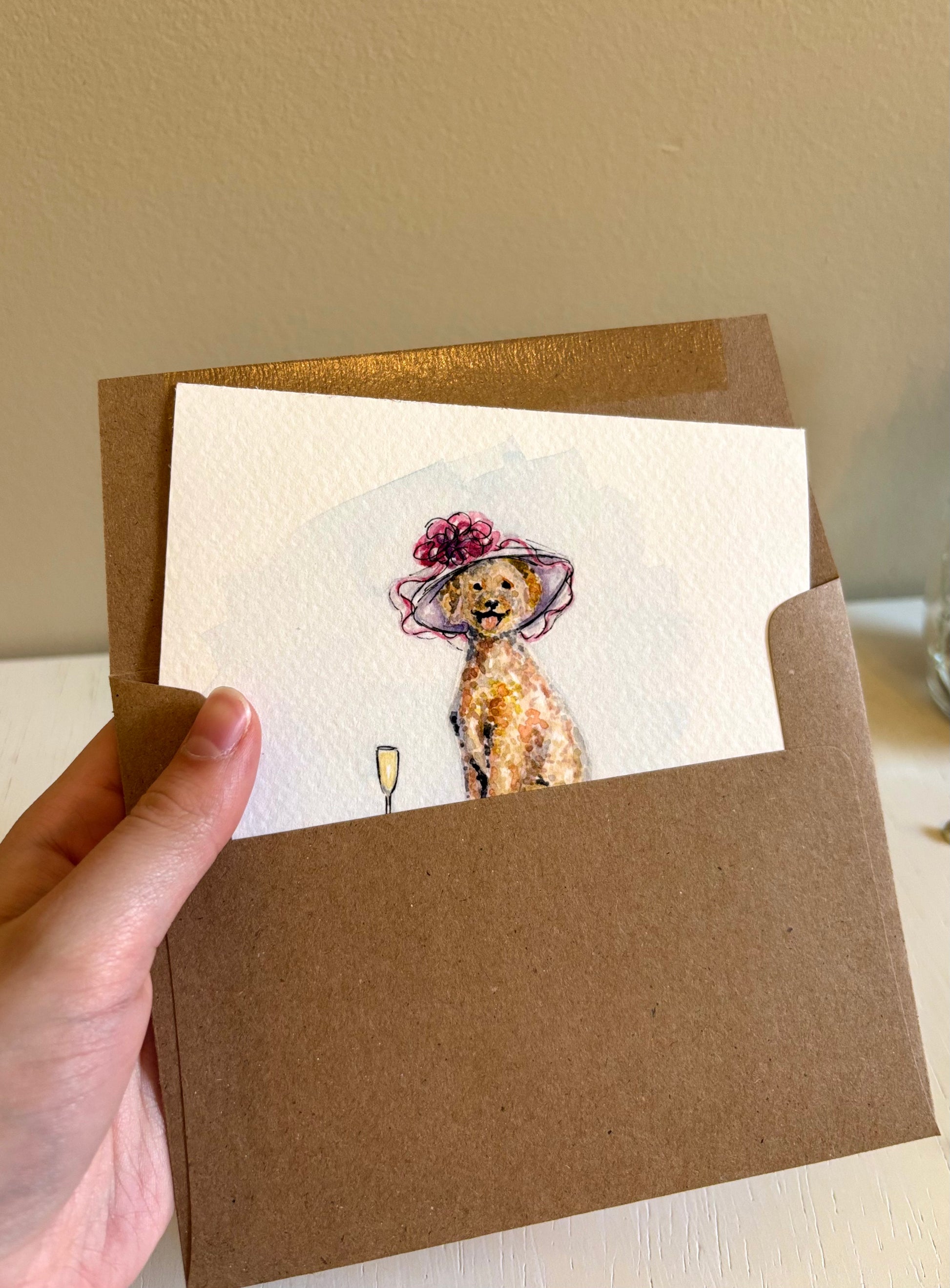 Hand holding a card with a dog illustration wearing a hat and holding a glass of wine, inside a brown envelope.