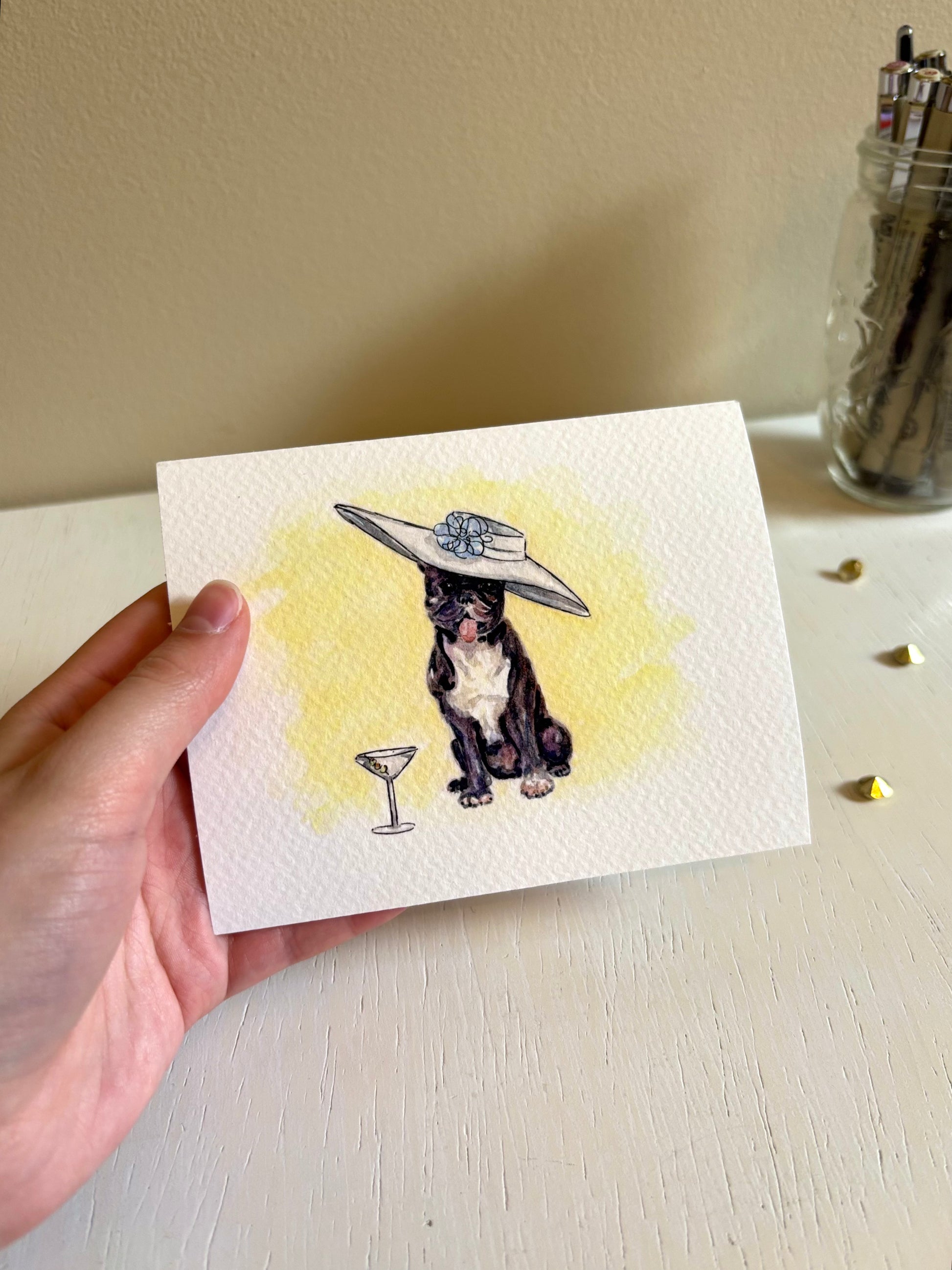 Hand holding a fine art print with a dog wearing a hat and holding a martini glass illustration.