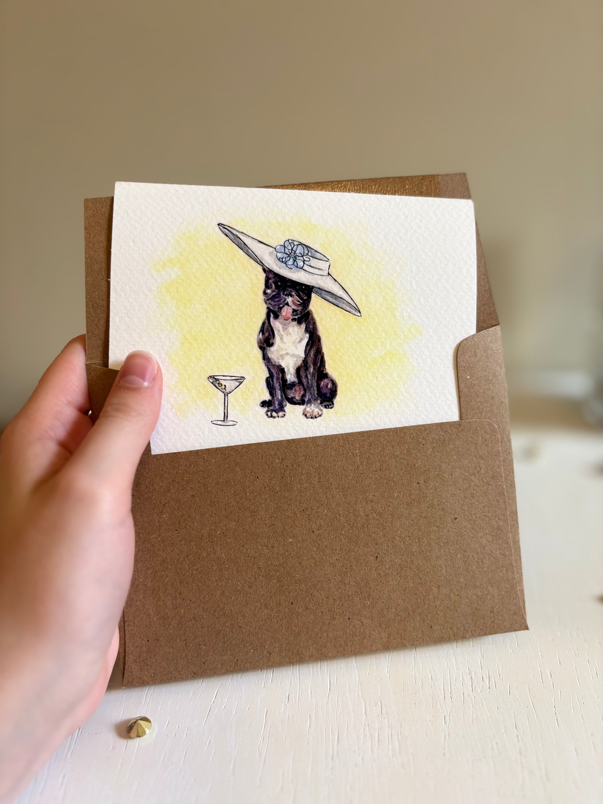 Hand holding a card with a dog illustration wearing a hat and a cocktail, on a brown envelope.