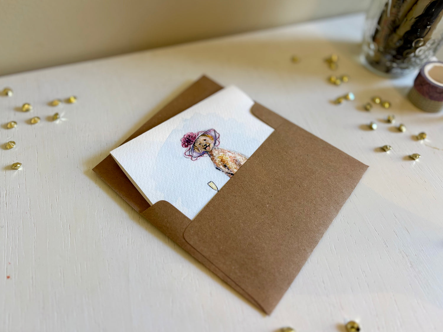 Watercolor greeting card partially in a brown envelope on a white surface with an aesthetic background.