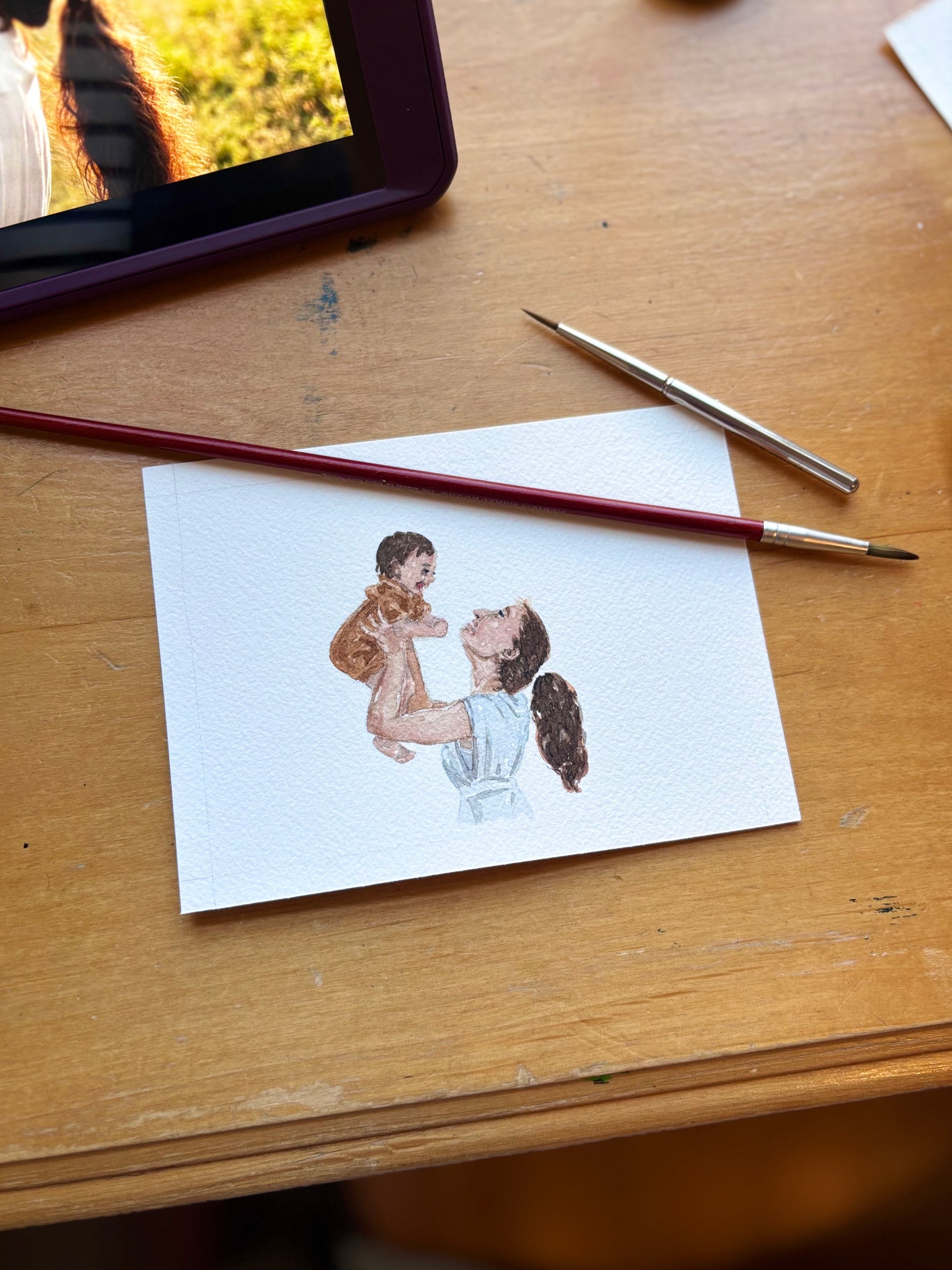Watercolor illustration of a woman holding a child on a piece of paper with two brushes on a wooden surface.