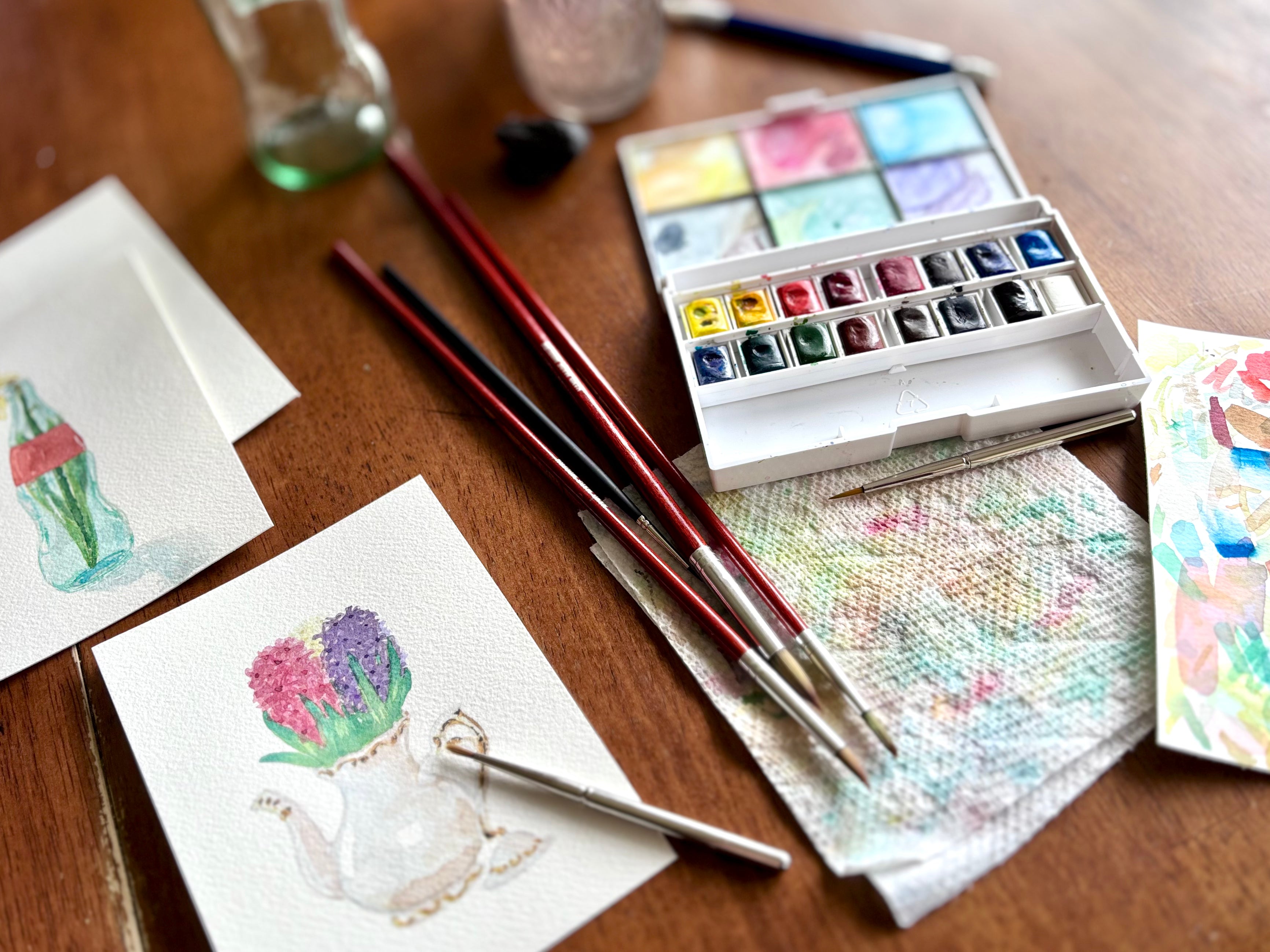 Greeting card watercolor illustrations on a brown table with brushes and paint palette. 