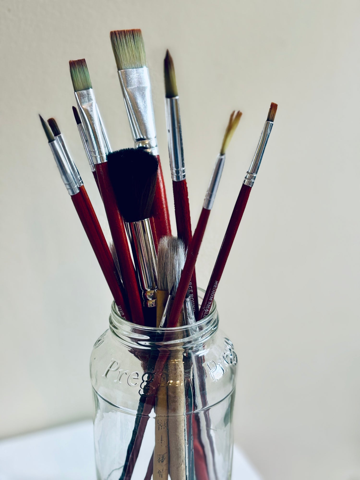 Set of watercolor paintbrushes in a clear glass jar on a plain, cream background.