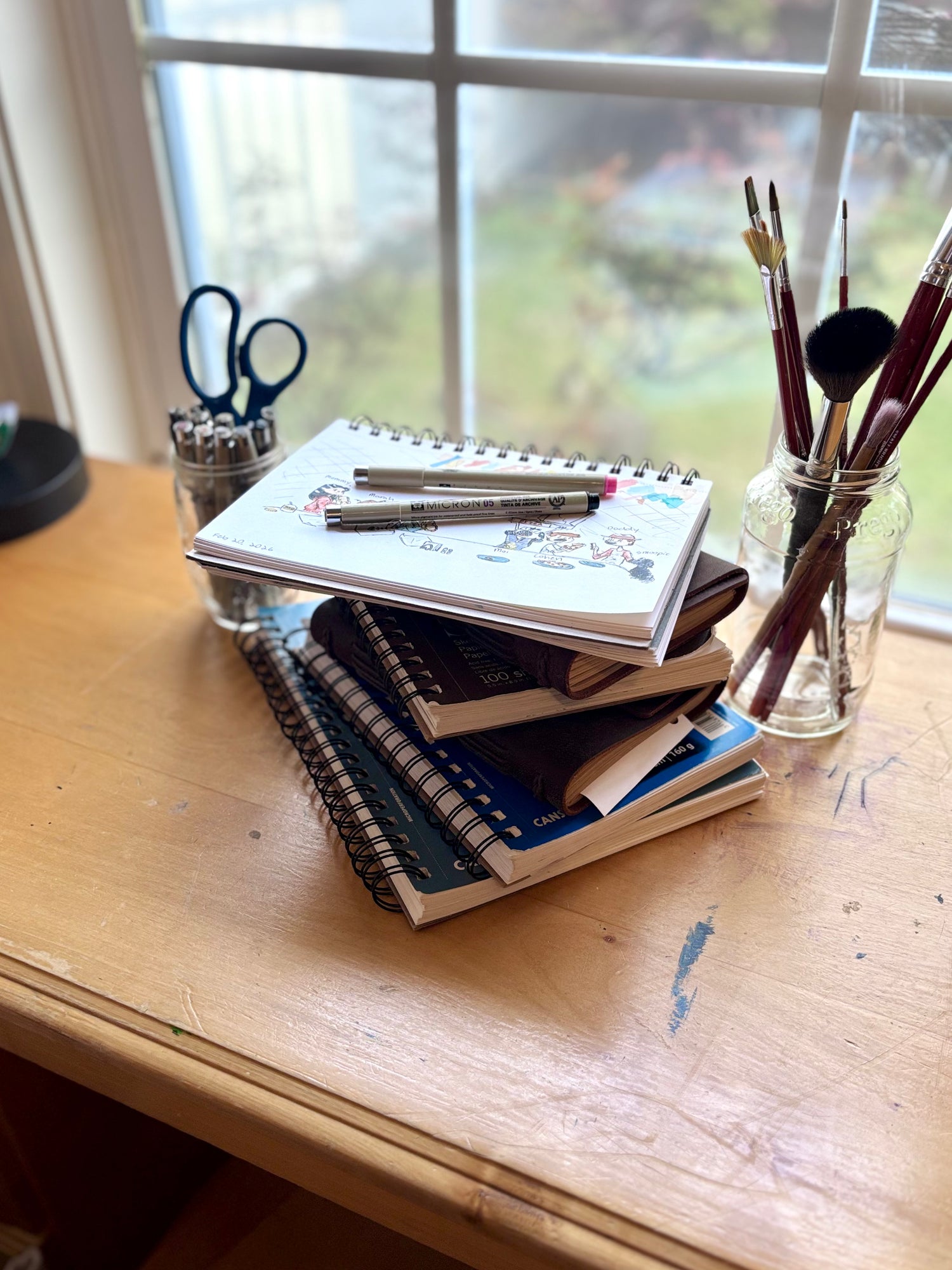 Stack of sketchbooks with pens, watercolor illustrations, and brushes on a wooden table near a window.